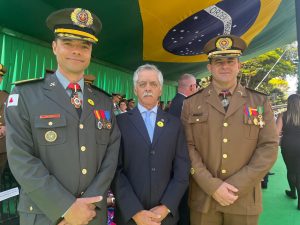 A imagem mostra três autoridades em pé, lado a lado, durante o desfile cívico-militar de 7 de setembro. O coronel Moisés Magalhães de Sousa, chefe do Estado-Maior do Corpo de Bombeiros Militar de Minas Gerais, vestindo farda oficial com boina; o coronel Sócrates Edgard dos Anjos, corregedor da Justiça Militar, usa terno preto com camisa azul; e o coronel Maurício José de Oliveira, chefe do Estado-Maior da Polícia Militar de Minas Gerais, veste farda marrom com boina. Ao fundo, aparece uma grande bandeira do Brasil compondo o cenário da solenidade