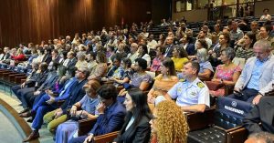 A imagem mostra a plateia da solenidade de celebração pelo 98º aniversário da UFMG, composta por diversas pessoas, entre homens e mulheres, vestidos em trajes formais. Os convidados estão sentados em poltronas pretas de auditório, e as paredes são de madeira 