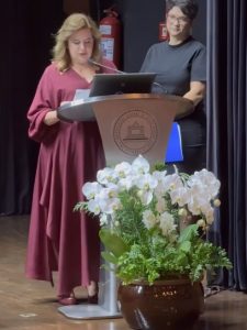 A imagem mostra a reitora da UFMG, Sandra Regina Goulart Almeida, em um púlpito metálico com o brasão da universidade, fazendo um discurso durante a solenidade de celebração pelo 98º aniversário da instituição. Ela usa vestido vinho de mangas longas e sapatos da mesma cor. Em primeiro plano, à frente do púlpito, há um arranjo de orquídeas brancas em vaso marrom
