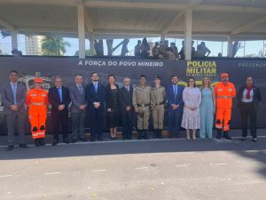 A imagem mostra 14 pessoas posando para foto durante solenidade da Polícia Militar de Minas Gerais, entre homens e mulheres civis e militares. Eles estão em ambiente externo, na frente de um palanque, com militares na parte superior da estrutura. Ao fundo há um painel com a inscrição “Polícia Militar de Minas Gerais – A Força do Povo Mineiro”, que compõe o cenário