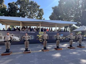 A imagem mostra o momento da passagem de comando com seis oficiais militares em pé, no pátio da APM. Eles estão enfileirados diante do comandante da APM e do palanque de autoridades