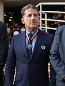 A imagem mostra um plano fechado do desembargador Fernando Antônio Nogueira Galvão da Rocha, que usa terno azul-marinho, camisa azul-clara e gravata azul-escura. No pescoço, ele está com a Medalha Alferes Tiradentes, a mais alta comenda da Polícia Militar de Minas Gerais. No lado esquerdo do seu peito, ele usa um broche. Ao fundo, parcialmente desfocado, percebem-se pessoas e a arquitetura de um prédio