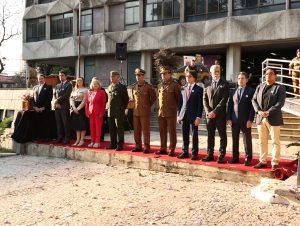 A imagem mostra a cerimônia da Medalha Alferes Tiradentes, com os homenageados em destaque, sendo 9 homens e duas mulheres. No centro, estão oficiais da Polícia Militar de Minas Gerais fardados. À direita, o desembargador Fernando Antônio Nogueira Galvão da Rocha, segundo da direita para a esquerda, veste terno escuro. Em frente ao grupo, um tapete vermelho e pétalas de flores caídas no chão. Ao fundo, um edifício de concreto aparente com grandes janelas, a luz do sol ilumina o ambiente
