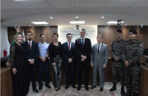 A imagem mostra um grupo de nove pessoas enfileiradas, entre autoridades civis e militares, durante a segunda palestra do Ciclo de Encontros Temáticos. Elas posam lado a lado no plenário do TJMMG, que tem como fundo uma parede de madeira com bandeiras à esquerda. 