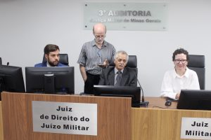 A imagem mostra três magistrados reunidos na 3ª Auditoria da Justiça Militar de Minas Gerais, sentados em frente a computadores sobre a bancada de madeira da sala de audiências da 3ª Auditoria de Justiça Militar. À esquerda da imagem está o juiz João Pedro Hoffert Monteiro de Lima, de terno azul; ao centro, o desembargador Sócrates Edgard dos Anjos, de terno cinza, camisa e gravata cinzas; e à direita a juíza Daniela de Freitas Marques, de óculos e camisa branca. De pé, atrás do desembargador, está um servidor de camisa listrada. Ao fundo, destaca-se a placa com a identificação da 3ª Auditoria.