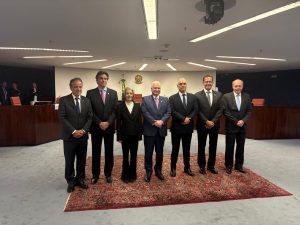 A imagem mostra sete pessoas em pé, lado a lado, em um ambiente formal, sobre um tapete vermelho no plenário do Supremo Tribunal Federal. Todos estão vestidos com roupas sociais escuras, em tons de preto e azul. O primeiro à esquerda da imagem é o desembargador James Ferreira Santos e posicionado ao centro está o ministro Luiz Edson Fachin. Ao fundo, aparece a bancada de madeira, cadeiras com estofado vermelho, a bandeira do Brasil e o brasão da República fixado na parede.