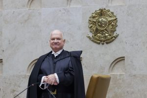 A imagem mostra o ministro Luiz Edson Fachin em pé, de toga preta, diante do plenário do Supremo Tribunal Federal, em Brasília. Ele sorri levemente enquanto posa para a foto, com as mãos apoiadas uma sobre a outra. Ao fundo, na parede de mármore claro, destaca-se o brasão da República em relevo dourado. À frente, aparece parcialmente uma cadeira marrom e os microfones do púlpito.