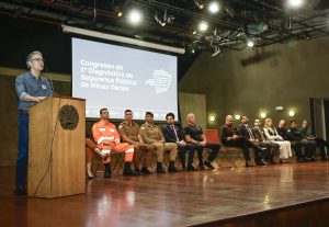 A imagem mostra o palco do auditório JK durante o “Congresso do 1º Diagnóstico de Segurança Pública de Minas Gerais”. À esquerda, o governador Romeu Zema fala ao microfone em um púlpito de madeira com o brasão do Estado de Minas Gerais. À direita, alinhadas em uma fila de cadeiras, estão diversas autoridades, entre homens e mulheres, representantes da forças de segurança e convidados civis. No fundo da imagem aparece um telão projetando o título do evento.