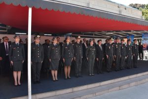 A imagem mostra autoridades civis e militares no palanque da cerimônia de formatura dos tenentes do Curso de Habilitação de Oficiais do CBMMG, entre elas, o juiz Marcos Luiz Nery Filho. Todos estão em posição de respeito, vestindo farda ou roupas formais e ocupando a extensão da tribuna coberta com tenda vermelha. Ao fundo, há banners institucionais que compõem o cenário