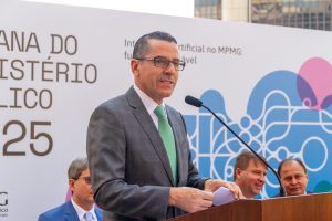 A imagem mostra o procurador-geral de Justiça, Paulo de Tarso Morais Filho, em destaque, falando ao microfone durante a abertura da Semana do Ministério Público. Ele usa óculos, veste terno cinza escuro com gravata verde e camisa branca, está posicionado atrás de um púlpito de madeira e segura papéis nas mãos