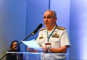A imagem mostra o diretor da Enajum e ministro do Superior Tribunal Militar (STM), Leonardo Puntel, em destaque, em um púlpito de acrílico falando ao microfone durante a abertura do Congresso Internacional de Direito Militar. Ele veste farda branca, usa óculos e segura alguns papéis enquanto discursa