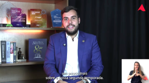 A imagem mostra trecho do vídeo de estreia da nova temporada da série "Você conhece o Direito Militar?". O juiz João Pedro Hoffert Monteiro de Lima está sentado em primeiro plano, de terno azul e camisa branca. O cenário tem um fundo preto e, à direita, uma estante com livros sobre Direito Militar e troféus conquistados pelo TJMMG. No canto inferior direito da imagem está a janela de libras com a intérprete vestindo camiseta preta