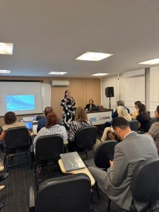 A imagem mostra a juíza em pé, falando ao microfone durante o Encontro de Cooperação Judiciária. Ela usa óculos e uma roupa estampada em preto e branco, posicionada à frente de uma plateia sentada em um auditório. 