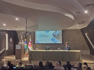 A imagem mostra o juiz em pé, falando ao púlpito durante o Encontro de Cooperação Judiciária. Ele veste terno cinza e gravata vermelha, e atrás dele estão três bandeiras. À direita, outro participante acompanha a apresentação sentado à mesa principal. No telão, é projetado o tema “Banco de Sentenças das Justiças Militares”, com os logotipos do STM, TJMMG, TJMRS e TJMSP. Ele fala para um público sentando no auditório