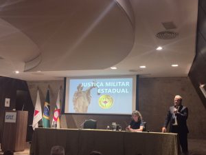 A imagem mostra o coronel em pé, falando ao microfone durante o Encontro de Cooperação Judiciária. Ele veste terno escuro e gravata azul, e ao fundo há uma apresentação projetada com o título “Justiça Militar Estadual” e o brasão do TJMMG. À mesa, uma participante acompanha a fala, enquanto as bandeiras estão dispostas ao lado do púlpito com o logotipo da Ejef