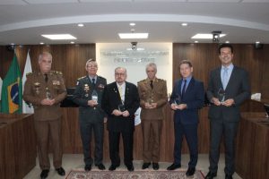 A imagem mostra seis homens em pé, lado a lado, em frente à tribuna da sala do Pleno do TJMMG. Todos estão vestidos com trajes formais civis e militares e seguram uma comenda nas mãos. Ao fundo, na parede, está escrito “Tribunal de Justiça Militar de Minas Gerais”, e, à esquerda, aparecem bandeiras