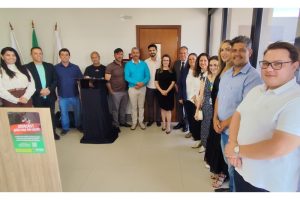 : A imagem mostra um grupo de 15 pessoas, entre homens e mulheres, reunidos em um ambiente interno durante a inauguração do PID em Papagaios. Dentre eles, está o desembargador James Ferreira Santos, vestindo terno escuro e gravata. Os participantes posam lado a lado em uma sala com paredes claras. Ao fundo há três bandeiras.