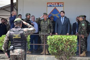 A imagem mostra uma cerimônia em área externa com a presença de militares e civis. Em primeiro plano, um militar usando uniforme camuflado e colete preto escrito “Polícia do Exército” faz continência. À frente, outros militares fardados retribuem a saudação, e o juiz Marcos Luiz Nery Filho está entre eles, vestindo terno e gravata. Ao fundo, aparece um letreiro com o brasão da 4ª Cia de Polícia do Exército 