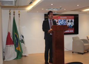 A imagem mostra o desembargador Fernando José Armando Ribeiro em pé atrás de um púlpito de madeira, falando ao microfone no auditório do TJMMG. Ele está vestindo terno e gravata. Ao fundo, há uma tela exibindo o tema do evento, e à esquerda da imagem estão três bandeiras.