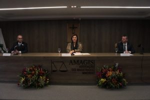 Imagem mostra três pessoas sentadas em uma mesa de debates na sede da Amagis. Os homens estão de terno e ao centro está uma mulher, de roupa com estampa de detalhes pretos. A mesa, bem como o painel de fundo, são de madeira