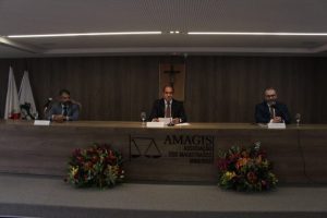 Imagem mostra três homens de terno sentados em uma mesa de debates na sede da Amagis. A mesa, bem como o painel de fundo, são de madeira