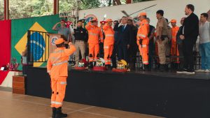 A imagem mostra um pátio com diversas autoridades em um palanque. Os bombeiros, vestidos de farda laranja, prestam continência ao desembargador coronel Osmar Duarte Marcelino, de terno escuro