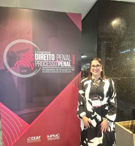 A imagem mostra a juíza em pé ao lado de um painel vertical escrito 1º Congresso de Direito Penal e Processo Penal do MPMG. Ela está de vestido estampado em branco e preto, usa óculos e tem os cabelos lisos soltos.