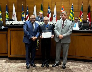 A imagem mostra três homens em pé, vestidos com ternos, posicionados à frente de uma mesa no Plenário Ministro Waldemar Zveiter. O corregedor da Justiça Militar de Minas Gerais, ao centro, segura um diploma com as mãos e usa uma medalha pendurada no pescoço. Ao fundo, há uma parede de madeira e bandeiras de diversos estados brasileiros