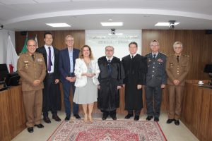 A imagem mostra oito pessoas em pé, sete homens vestidos formalmente com ternos, e uma mulher usando vestido, uma ao lado da outra, no Plenário do TJMMG. Ao fundo na parede, está escrito "Tribunal de Justiça Militar de Minas Gerais” e, à esquerda, aparecem bandeiras. Da esquerda para a direita, estão: o desembargador James Ferreira Santos; o desembargador Fernando Armando; o professor Alessandro Fernandes Moreira, vice-reitor da UFMG; a professora Sandra Regina Goulart Almeida, reitora da UFMG; o presidente Jadir Silva; o desembargador Fernando Galvão; o desembargador Osmar Duarte Marcelino e o desembargador Sócrates Edgard dos Anjos. 