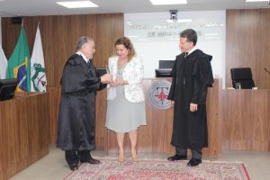A imagem mostra os desembargadores vestindo toga e entregando a comenda a professora Sandra Regina Goulart Almeida que usa um vestido cinza e um casaco branco florido. Eles estão no Tribunal Pleno, em que há três bandeiras ao fundo e uma parede de madeira. 