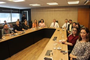 Na Sala Diamante do TJMMG, 14 pessoas estão sentadas ao redor de uma grande mesa de reunião. O grupo é composto por homens e mulheres, vestidos formalmente com trajes sociais. O ambiente tem paredes revestidas em tom amadeirado e preto, com uma ampla janela ao fundo