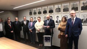 A imagem mostra um grupo de autoridades em um ambiente interno com paredes brancas apresentando os retratos da galeria. À direita da imagem está o juiz Luiz Carlos Rezende e Santos usando terno azul e óculos. À esquerda está o desembargador Jadir Silva usando terno preto e gravata vermelha. Um dos participantes faz o uso da palavra atrás do púlpito da Amagis