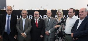 A imagem mostra seis homens e uma mulher, posando para foto em pé. O grupo está em frente a uma mesa de autoridades, em ambiente interno iluminado. Todos usam trajes sociais e a mulher um vestido floral longo. Ao centro está o desembargador Jadir Silva usando terno preto com gravata vermelha e óculos. Ao seu lado direito está o homenageado, juiz aposentado Décio de Carvalho Mitre, vestindo terno cinza e gravata listrada