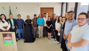 A imagem mostra um grupo de 15 pessoas, entre homens e mulheres, reunidos em um ambiente interno durante a inauguração do PID em Papagaios. Dentre eles, está o desembargador James Ferreira Santos, vestindo terno escuro e gravata. Os participantes posam lado a lado em uma sala com paredes claras. Ao fundo há três bandeiras.