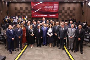 A imagem mostra um grupo de cerca de 30 pessoas, entre homens e mulheres, no Auditório do Pleno do TJMG. Todos estão de pé, lado a lado, de ternos e trajes formais. Ao fundo, há um grande painel eletrônico vermelho escrito “XVIII Consepre”. À esquerda, no fundo do auditório, aparecem diversas bandeiras dispostas em fileira.