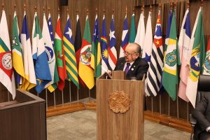 A imagem mostra o desembargador Jadir Silva, que veste terno preto e gravata roxa, discursando em uma tribuna de madeira no Auditório de Pleno do TJMG. Ao fundo, há uma fileira de bandeiras dispostas lado a lado.