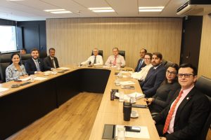 Na Sala Diamante do TJMMG, 11 pessoas estão sentadas ao redor de uma grande mesa de reunião. O grupo é composto por homens e mulheres, vestidos formalmente com trajes sociais, todos atentos à uma apresentação. Ao centro do grupo está o corregedor da Justiça Militar, que veste camisa clara e gravata escura. O ambiente tem paredes revestidas em tom amadeirado e preto