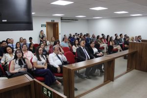 A imagem mostra o público reunido no Plenário do TJMMG durante a capacitação, com dezenas de participantes ocupando as fileiras de cadeiras vermelhas e pretas. Entre os presentes, homens e mulheres acompanham a apresentação