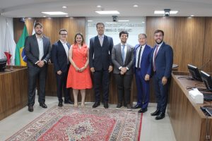 A imagem mostra um grupo de sete pessoas, entre desembargadores, juízes e o palestrante Caio Perona, reunidos no Plenário do TJMMG. Seis são homens, vestidos de terno, e a mulher na imagem é a juíza Carolina Aleixo Benetti de Oliveira Rodrigues, de vestido laranja. Ao fundo, é possível ver o painel institucional do Tribunal, com paredes revestidas em madeira e bandeiras do Brasil e de Minas Gerais