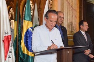 : A imagem mostra três autoridades do Judiciário durante a assinatura da Carta de Belo Horizonte. Em primeiro plano, o vice-presidente do TJMMG aparece assinando o documento, vestindo camisa social clara e segurando uma caneta sobre uma tribuna de madeira. À direita, estão o presidente do Consepre e o presidente do TJMG, ambos de terno. Ao fundo, bandeiras oficiais compõem o cenário, em um ambiente interno