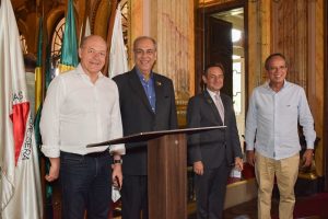 A imagem mostra quatro homens em pé atrás de uma tribuna em um ambiente com paredes claras e bandeiras ao fundo. Da esquerda para a direita: O presidente do TJMSP veste camisa social branca de manga comprida e calça jeans escura. O presidente do TJRO veste um terno preto com uma camisa azul. O presidente do TJMG veste um terno cinza-escuro com uma camisa clara e uma gravata listrada. O vice-presidente do TJMMG veste uma camisa social azul clara de manga comprida e calça bege.