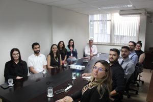 A imagem mostra um grupo de dez pessoas em uma sala de reuniões, sentados em torno de uma grande mesa de reunião, participando do programa Ambientar. Eles usam trajes formais. Há uma janela com cortina clara no fundo