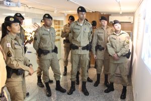 A imagem mostra um grupo de nove alunos do Curso de Formação de Soldados no Memorial da Justiça Militar. Eles estão uniformizados, com fardas bege e boinas pretas, observando o conteúdo exposto em uma das paredes do espaço. 