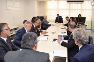 A imagem mostra os participantes da reunião sentados ao redor de uma mesa retangular em uma sala de reuniões com amplas persianas ao fundo. Entre eles está o desembargador James Ferreira Santos. Todos os presentes vestem trajes formais, com papéis, pastas e celulares dispostos sobre a mesa