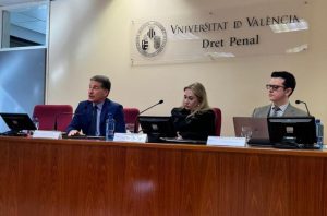 A imagem mostra três participantes sentados à mesa. À esquerda, está o desembargador de terno azul-marinho, camisa clara e gravata escura, falando ao microfone. Ao centro, a professora tem cabelos loiros e roupa preta. À direita, está o juiz usando terno claro, camisa social e gravata estampada. Todos estão posicionados atrás de computadores, em um auditório de paredes claras e com um letreiro da Universidade de Valência ao fundo