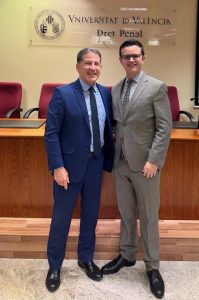 A imagem mostra duas pessoas posando lado a lado em um auditório. À esquerda, o desembargador veste terno azul-marinho, camisa clara e gravata escura. À direita, o juiz aparece de terno cinza, camisa social clara e gravata estampada. Ambos estão em frente à mesa principal, que possui cadeiras vermelhas e, ao fundo, um letreiro da Universidade de Valência 