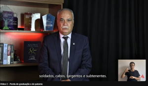 A imagem mostra o desembargador Rúbio Paulino Coelho, trajando terno escuro e camisa clara. Ao fundo, há uma prateleira com troféus da Justiça Militar e livros sobre Direito Militar. No canto direito da imagem está a janela de Libras, com a intérprete vestida de camiseta preta.