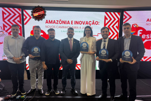 A imagem mostra um grupo de sete pessoas, sendo seis homens e uma mulher, premiados durante o Prêmio Inovação 2025. Eles estão em cima de um palco, em frente a um telão colorido. O penúltimo à direita é o juiz de Direito João Pedro Hoffert Monteiro de Lima, de terno preto, camisa branca e gravata preta com detalhes brancos. Ele sorri para a foto e segura o troféu em formato arredondado, de cor azul e preta.