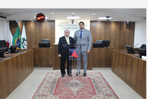A imagem mostra o presidente do TJMMG usando terno preto e gravata amarela. Ao seu lado está o juiz titular da 5ª AJME vestindo terno cinza e gravata azul. Eles estão em cima de um tapete vermelho e atrás de uma tribuna de vidro com a logo do TJMMG. Ao fundo há três bandeiras e uma parede de madeira.