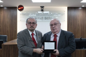 A imagem mostra o presidente do TJMMG usando terno preto e gravata vermelha. Ao seu lado está o juiz aposentado João Libério da Cunha vestindo terno cinza e gravata vermelha. Eles estão no Tribunal Pleno do TJMMG. Ao fundo há uma parede de madeira.